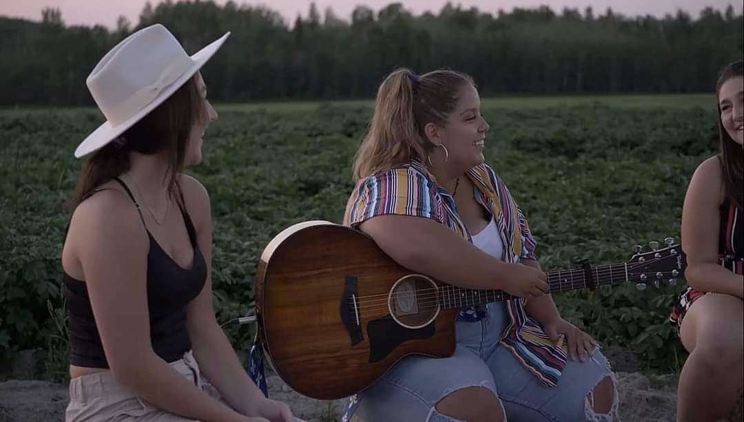 On voit Juliette assise entre deux de ses amies. Julienne tient une guitare dans ses mains. L'amie à sa gauche porte un chapeau blanc. Derrière elles, un champ bien vert.